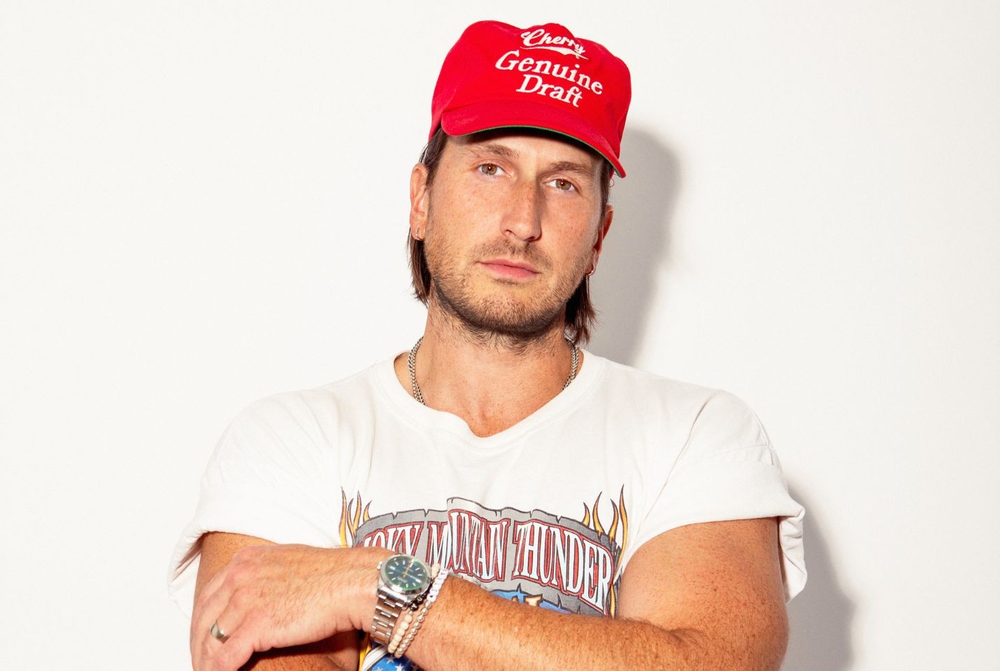 A man in a white graphic tshirt and red cap stood in front of a white wall